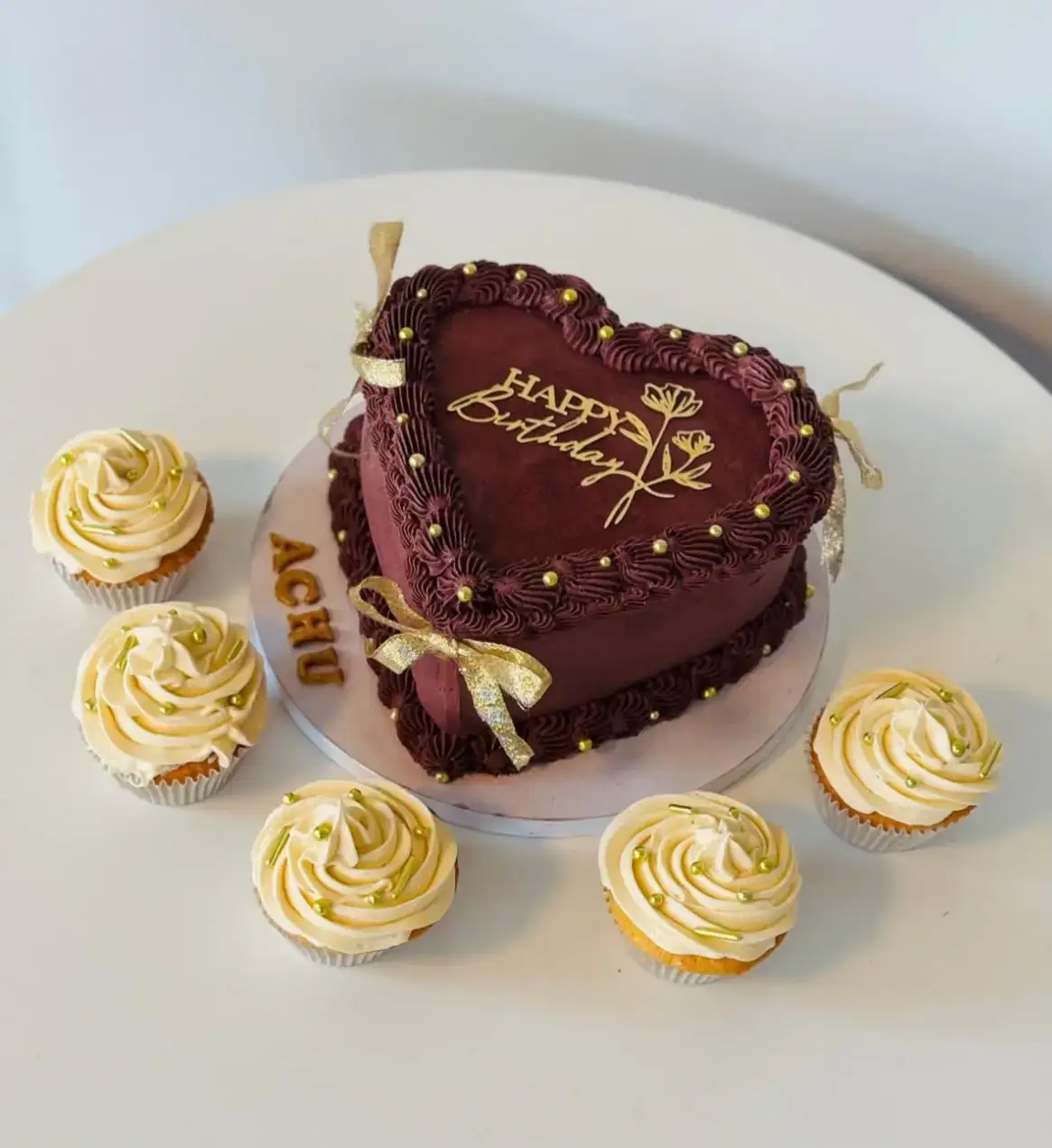 Bordeauxrote Herztorte mit goldener Geburtstagsaufschrift, umgeben von Cupcakes mit hellen Cremeblüten.